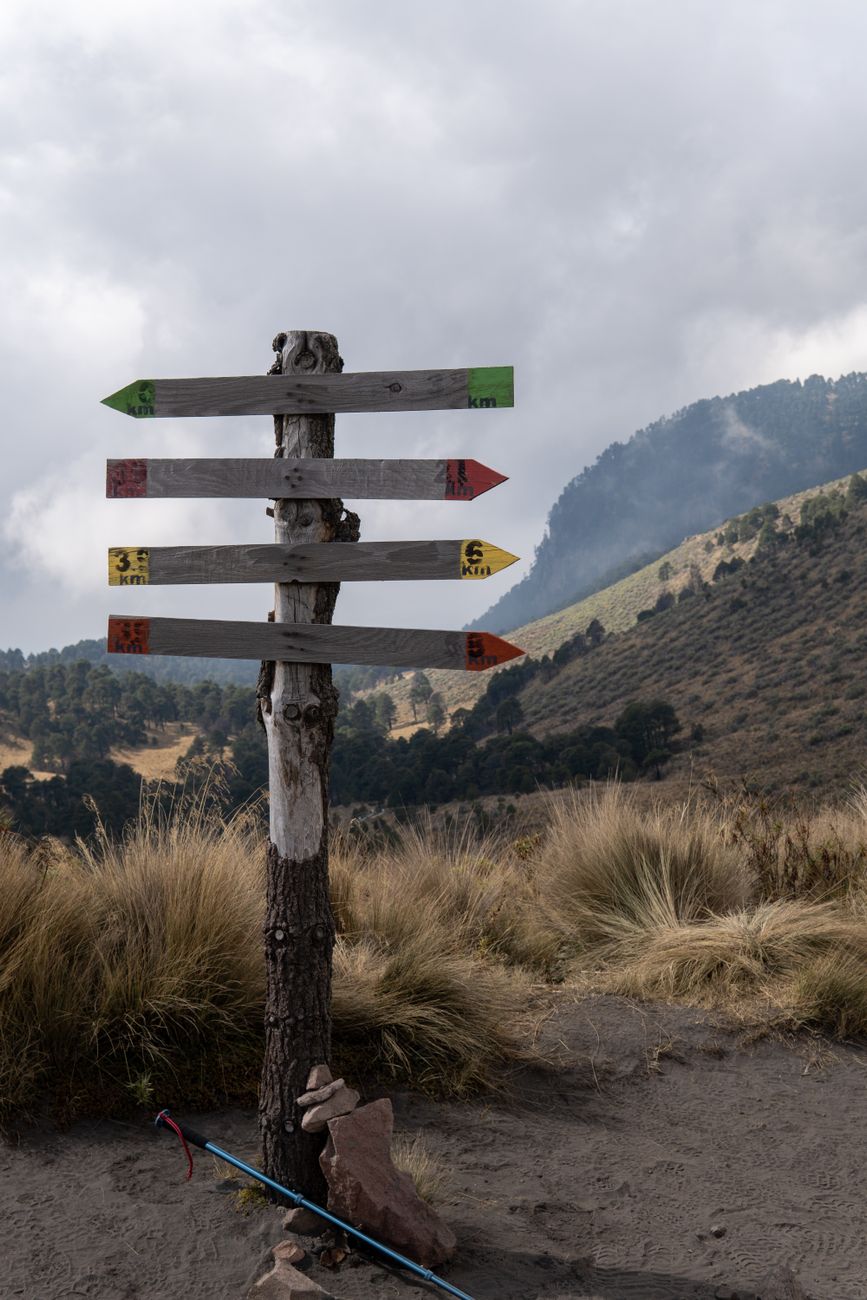 A post with four trail signs pointing in different directions. Tree-covered hills are in the background.